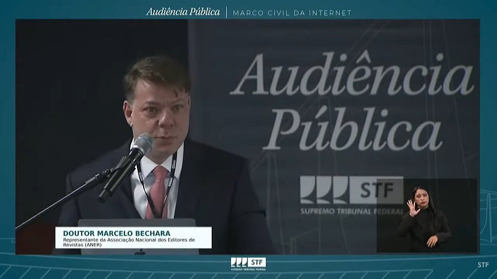 Homem branco de terno escuro, Marcelo Bechara fala à frente de tela escura da Audiência Pública com logotipo do STF