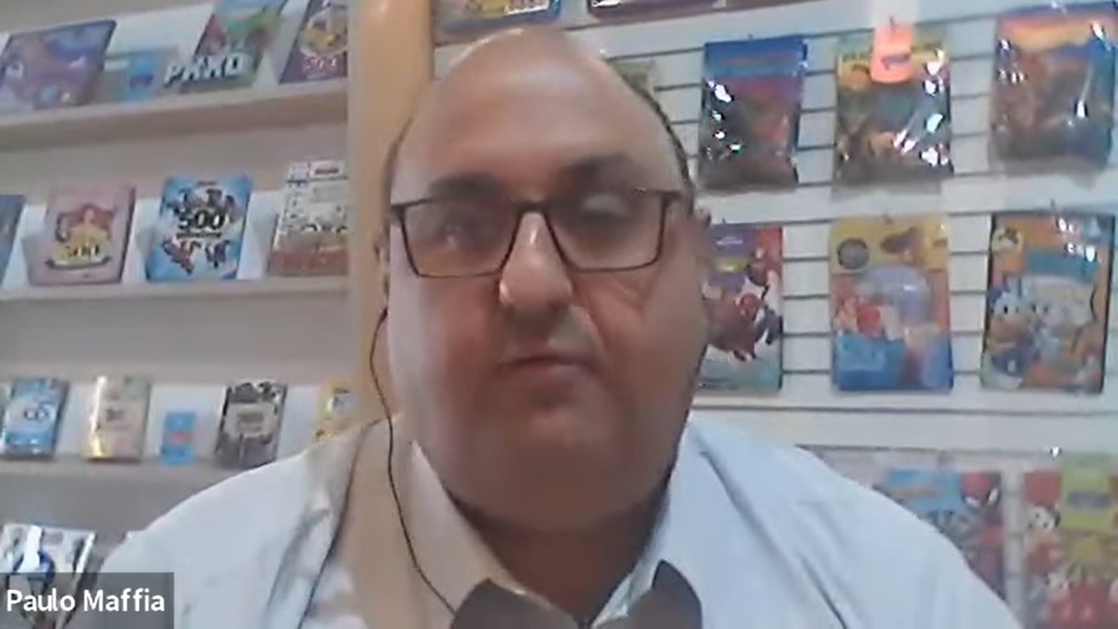 Homem branco de óculos quadrados e calvo, Paulo Maffia aparece em frente a uma parede que exibe várias revistas de histórias em quadrinhos hq coloridas