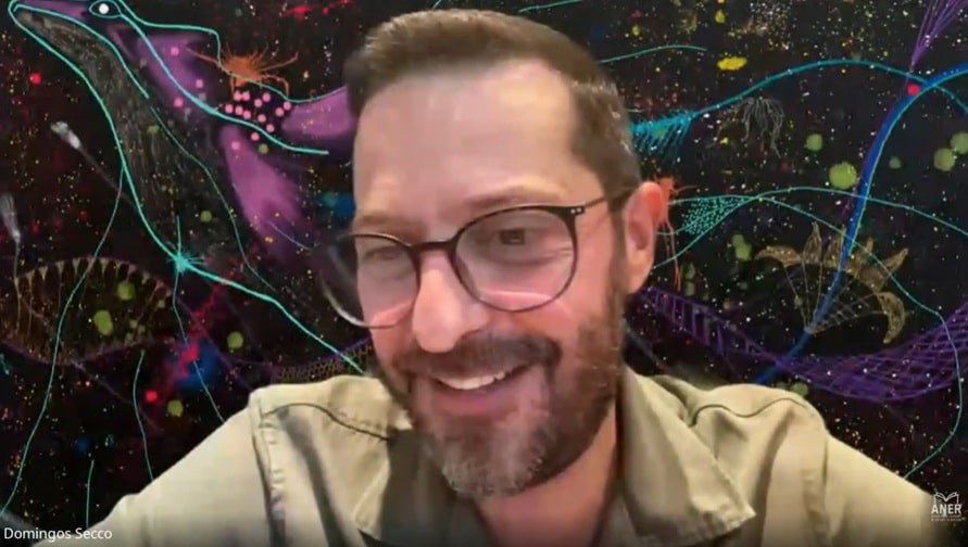 Domingos Secco da Alright homem branco, de cabelos, barba e bigode castanhos, vestndo blusa cáqui de gola e usando óculos de armação escura aparece em fundo colorido falando sobre as tramas do invisível.